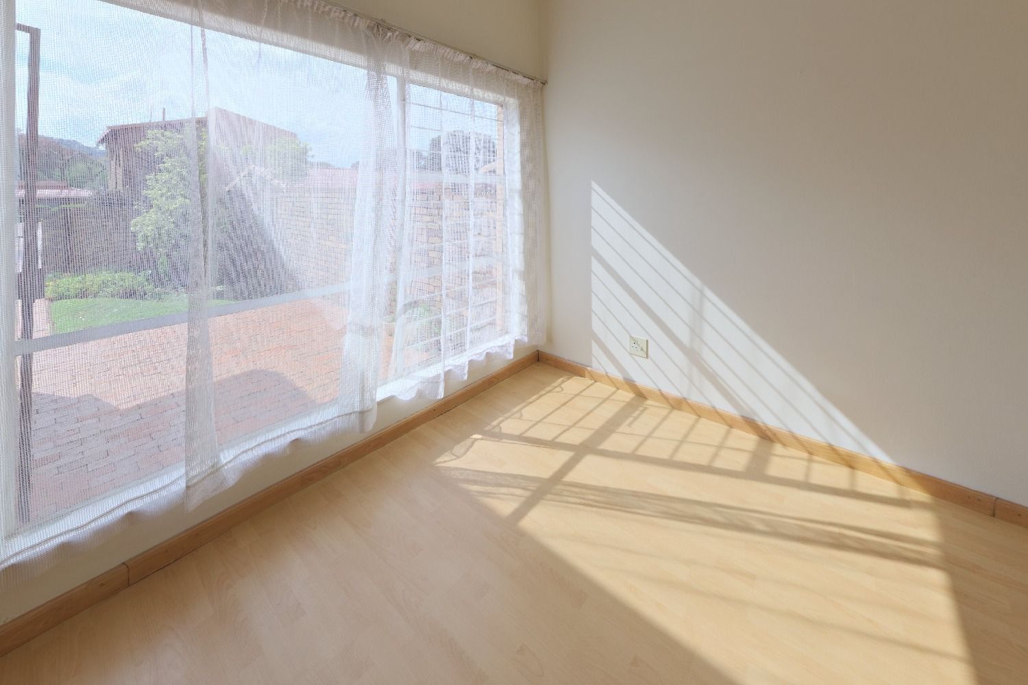 Original listing photo of an empty living room with a large window, sheer curtains and a light wood floor, before virtual staging