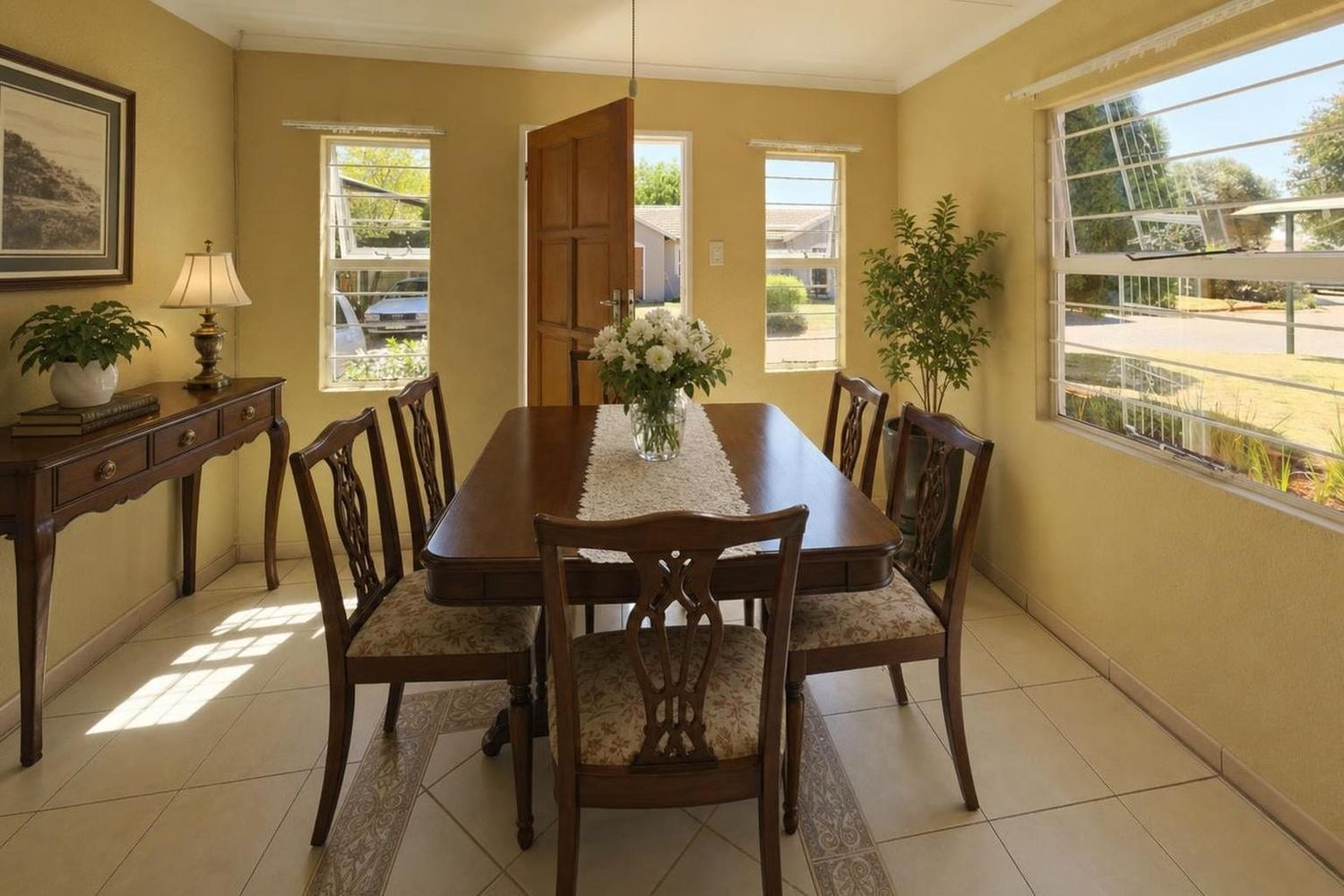 The same room virtually staged as a traditional dining room with a dark wood table, six carved chairs, a console table with a lamp and a vase of white flowers