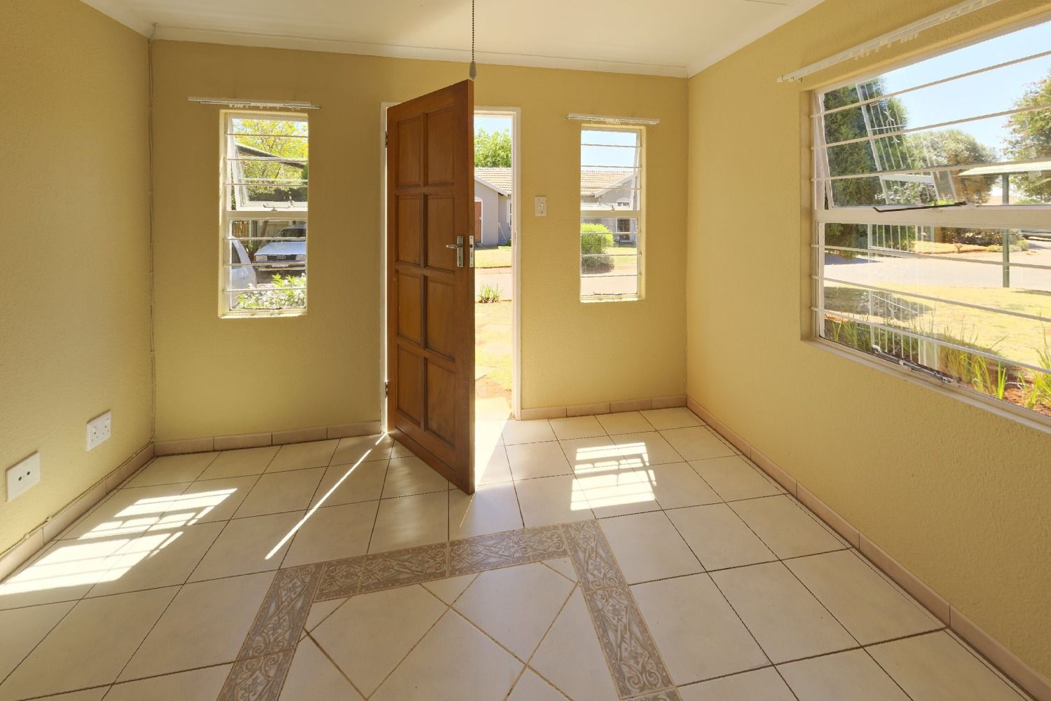 Empty front room with yellow walls, a tiled floor, a wooden front door and three windows, before being virtually staged as a dining room