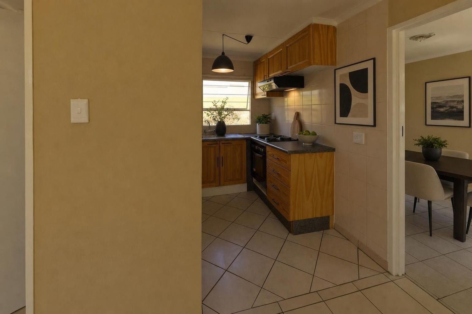The same kitchen virtually staged with a black pendant light, abstract art, plants and counter accessories, and a dining table visible in the next room